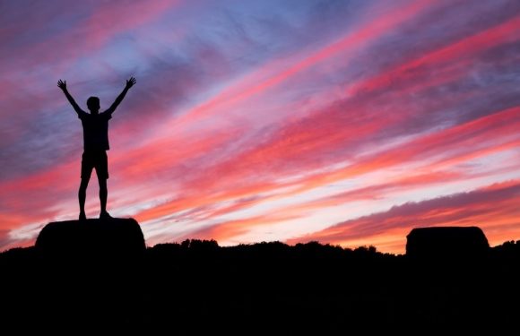 A person standing on a rock with arms raised, silhouetted against a vibrant sunset sky with pink, orange, and purple clouds, and distant landscape.