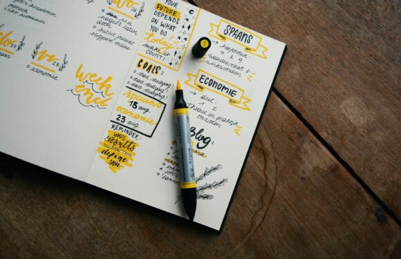 A white notebook open on a wooden table, filled with handwritten notes and highlights in yellow, along with a black and yellow marker, a yellow pushpin, and a small black binder clip.