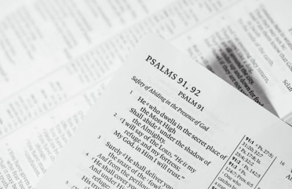 A close-up of a printed page featuring Psalms 91: 1-2 with other pages and a phone charger partially visible in the background.