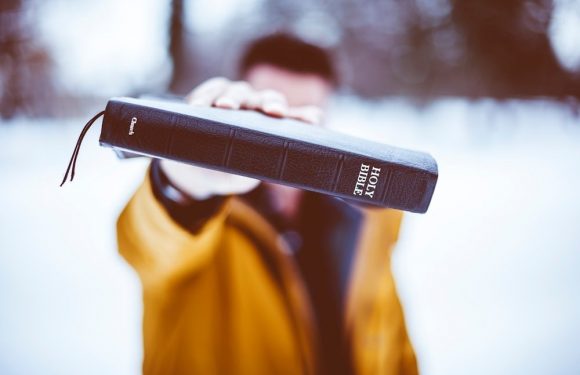 Person holding a Holy Bible outdoors, wearing a yellow jacket, with blurred snow and trees in the background.