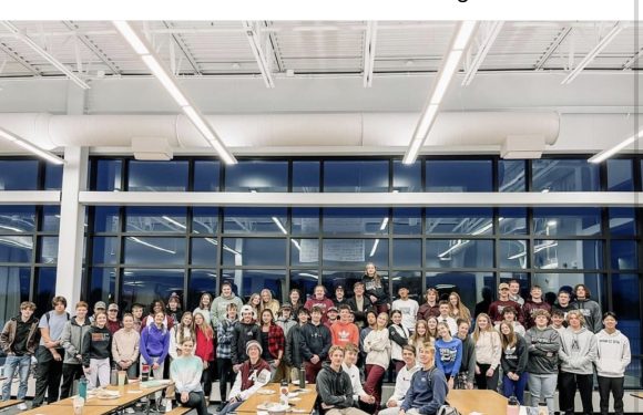 Group of students gathered in a large, well-lit cafeteria with food boxes and pizza along with a large banner on the wall.