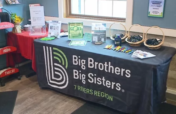 A table covered with a black cloth featuring the Big Brothers Big Sisters logo and "7 Rivers Region" text, displaying flyers, promotional items, and baskets with headphones.
