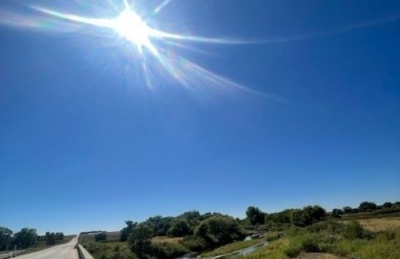 Bright sun in a clear blue sky over a rural landscape with a small river, grassy fields, and a bridge crossing the water.