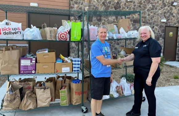 A man and woman shake hands in front of a metal rack filled with paper grocery bags and plastic bags outside a building with a stone wall.