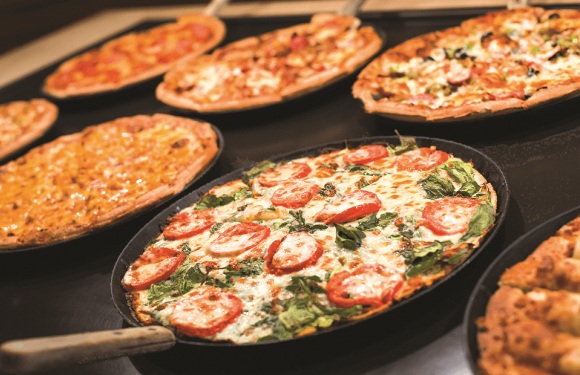 Several pizzas with different toppings are displayed on black trays, including a pepperoni and vegetable pizza in the foreground.