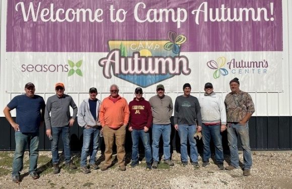 Ten people stand in front of a large banner that says "Welcome to Camp Autumn!" The group is outdoors on a gravel surface, and the banner promotes Camp Autumn.