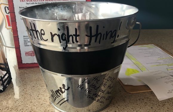 A metal bucket with black and handwritten text, placed on a countertop, with a sign behind it that mentions gluten sensitivity and pizza options.