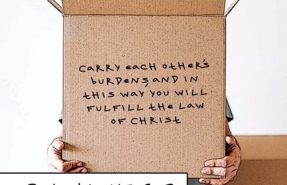 Person holding a cardboard sign with a handwritten Bible verse from Galatians 6:2. The person is partially obscured behind the sign, with hands visible.