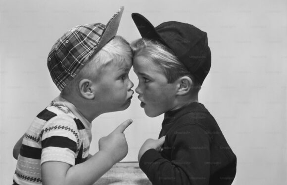 Two young boys face each other closely, with one finger raised as if making a point. They are wearing baseball caps and casual clothes, appearing to be in a playful argument.