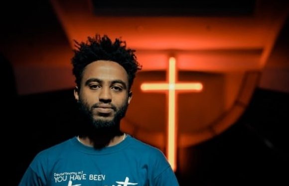 A young man with curly hair and a beard stands in front of a neon cross, wearing a blue T-shirt with white text about being set apart and chosen.