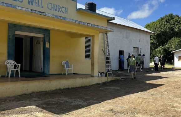 A group of people work on painting or renovating a small church building with a yellow front and white back, under a partly cloudy sky, with a dirt ground in the foreground.