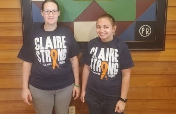Two girls in matching dark t-shirts with "Claire Strong" and a yellow ribbon stand indoors in front of a colorful geometric artwork mounted on wood-paneled wall.