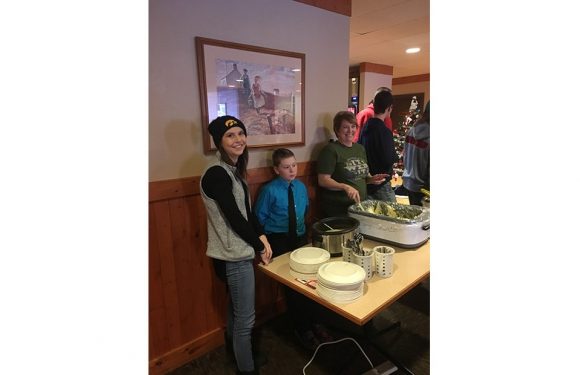 A woman in a green shirt serving food at a buffet table, with two children beside her, in a cozy restaurant decorated for Christmas.
