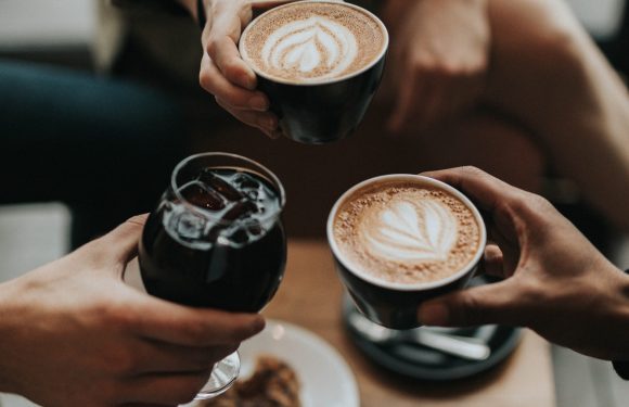 Three people are toasting with coffee cups and a glass of iced drink over a wooden table, which also has a plate with a cookie, empty glasses, and a spoon.