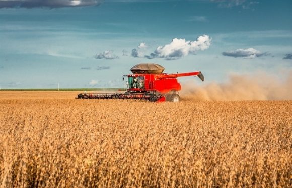 A large red combine harvester is operating in a wheat field under a partly cloudy sky, kicking up dust as it collects the crop.