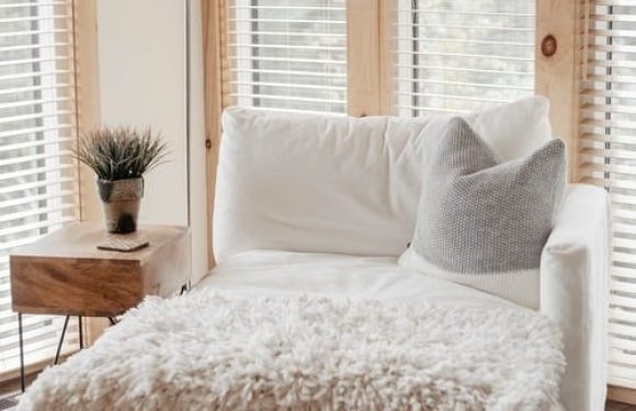 A cozy white armchair with a plush throw blanket and a gray pillow, next to a small wooden side table with a plant, all set near large windows with wooden blinds.
