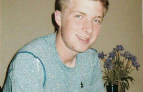 A young man with short, light brown hair, smiling and wearing a gray T-shirt, sitting near a vase of purple flowers against a neutral background.