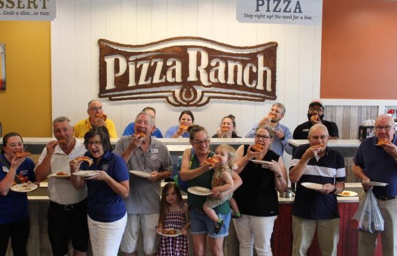 A group of casually dressed people, including children, enjoying slices of pizza in front of a "Pizza Ranch" sign inside a restaurant.