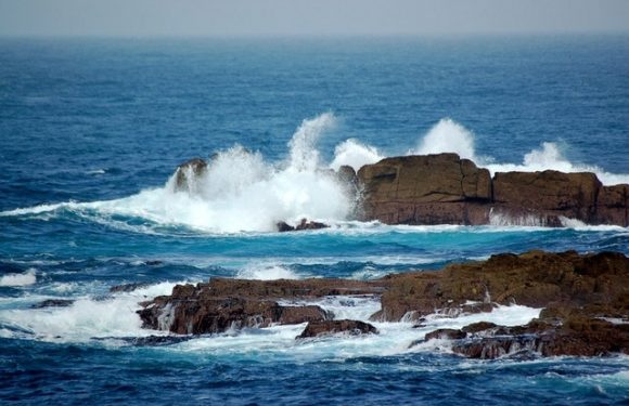 Waves crashing against rocky shoreline with the open ocean in the background, under a clear sky.