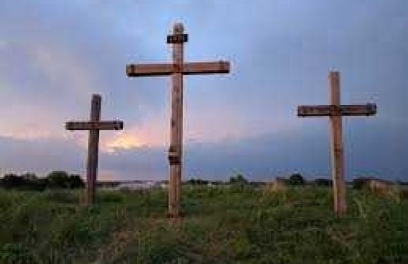 Three large wooden crosses stand on a grassy hill beneath a cloudy sky. Low horizon with some distant landscape visible.