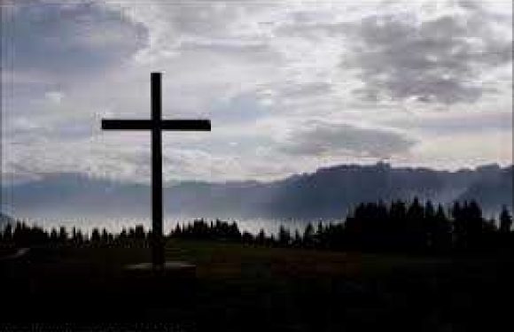 A large black cross stands outdoors against a cloudy sky with mountain silhouettes in the distance and a forested landscape below.