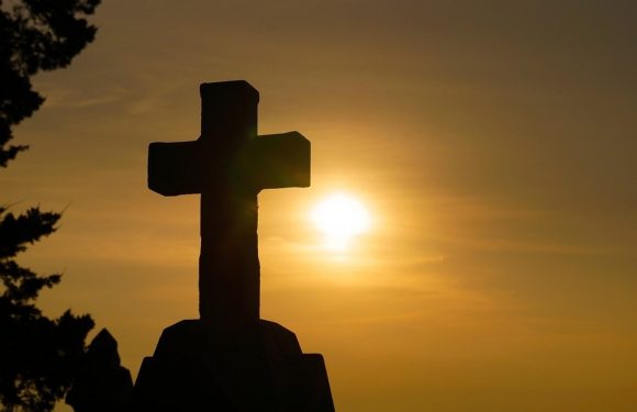 Silhouetted cross monument against a bright sunset or sunrise sky. A tree is partially visible on the left side of the image.