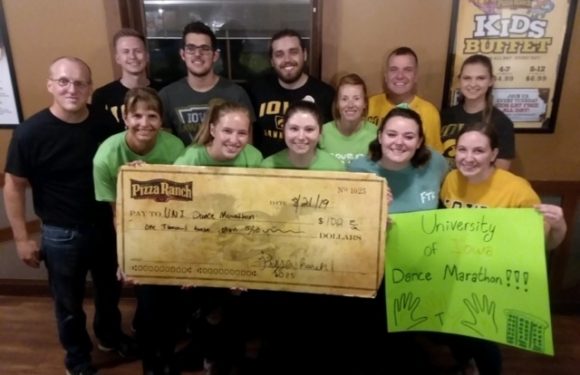 Group of people, some in green and yellow shirts, holding a large check and a bright green sign, celebrating a university dance marathon achievement indoors.