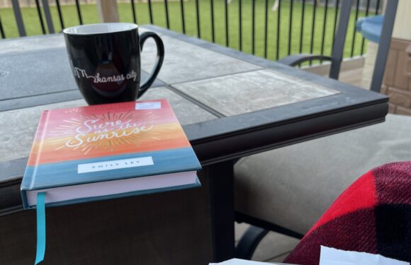 A black mug with "Kansas City" written on it, a colorful book titled "Sunshine and Sunsets" by Emily Lex, and an open Bible on a patio table with a wooden fence and grassy yard in the background.