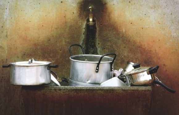 Several metal pots and pans rest on a rusty stove with a dark, sooty wall in the background. The scene appears to be in a kitchen or outdoor cooking area.
