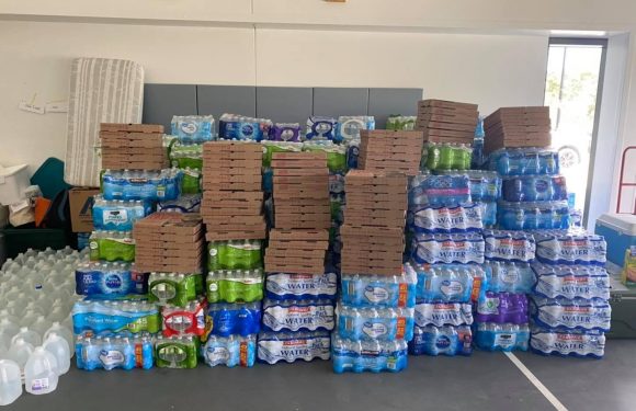 Stacks of bottled water and pizza boxes arranged on a table in a bright room with a window in the background. Two people are partially visible behind the items.