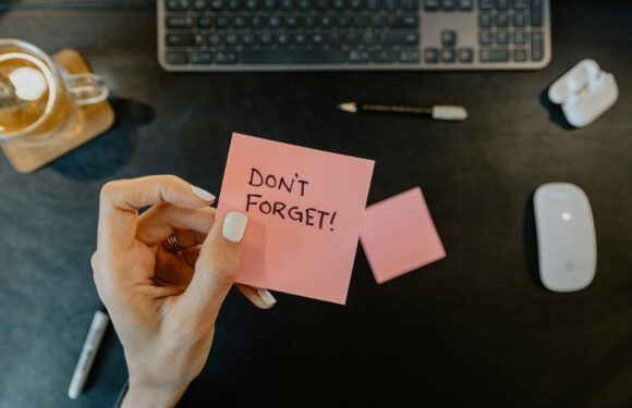 Hand holding a pink sticky note with "DON'T FORGET!" written on it, on a dark desk with a keyboard, mouse, pen, and a memory card nearby.