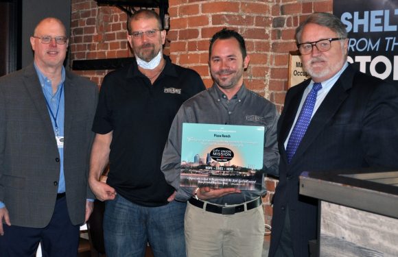 Four men standing in front of a brick wall, with one holding a sign; three men are dressed in suits or business casual, one in a Pizza Ranch uniform.