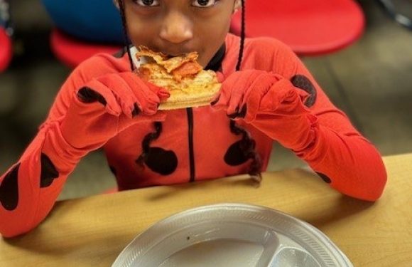 A girl with braided hair eats pizza while sitting at a table, wearing a red jacket with black heart shapes, and a divided metal plate is in front of her.