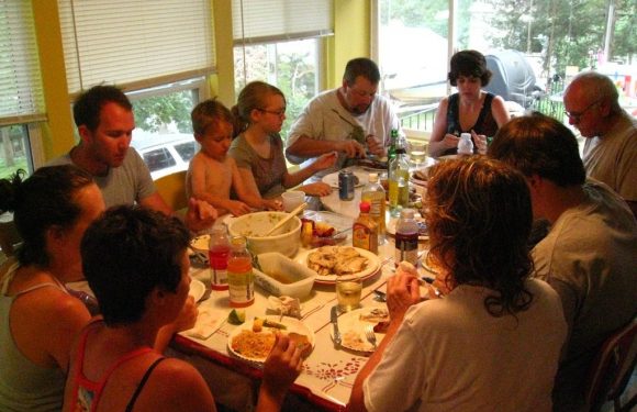 A group of people, including children and adults, gathered around a table enjoying a meal indoors, with windows showing greenery outside.