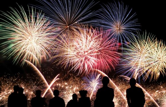 Six silhouetted people are watching a vibrant fireworks display against a black night sky, with multicolored explosions illuminating the scene.