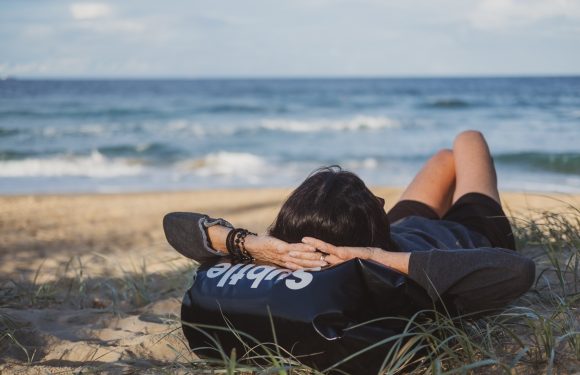 Person lying on the beach with hands behind head, wearing a dark jacket with "Staff" written on it, enjoying the sandy shore and ocean view.