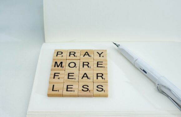 Scrabble tiles spell out "PRAY MORE FEAR LESS" on white paper with a white pen beside them, inside a minimalistic, light-colored setting.