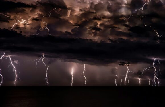 Dark stormy sky with multiple lightning strikes over a body of water, illuminated by flashes, with heavy clouds above.