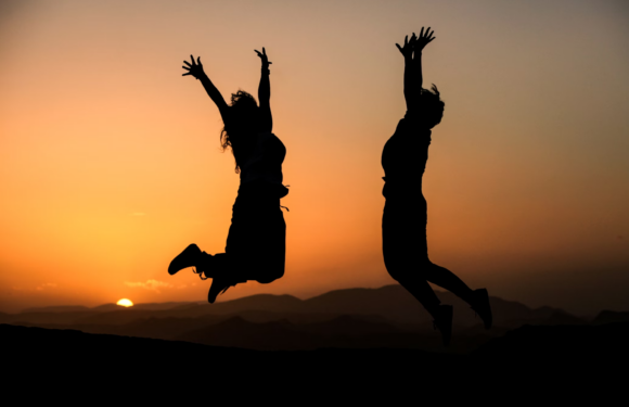 Two people are jumping in the air against a sunset with a mountainous landscape in the background. Their silhouettes are visible, and they appear to be joyful.