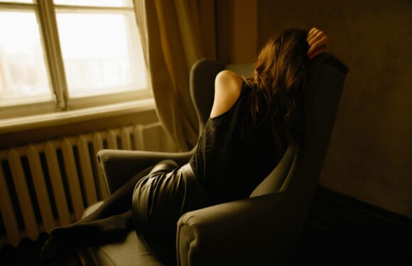 A woman with long, wavy hair sits in a dark armchair near a window with soft, natural light. She wears a black outfit with shoulder cutouts and appears contemplative.