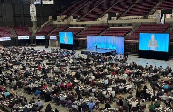 A large indoor auditorium filled with people sitting at tables, facing a stage with a speaker and two large screens displaying the speaker.
