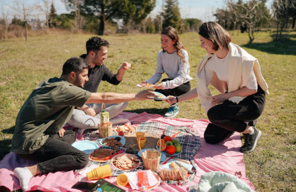 A group of four people sharing a picnic on a blanket outdoors, passing food and smiling, with a variety of snacks like bread, fruit, and cookies on the blanket.