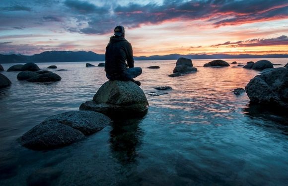 A person sits on a large rock in calm water during a colorful sunset or sunrise with dramatic clouds, mountains in the distance, and a peaceful atmosphere.