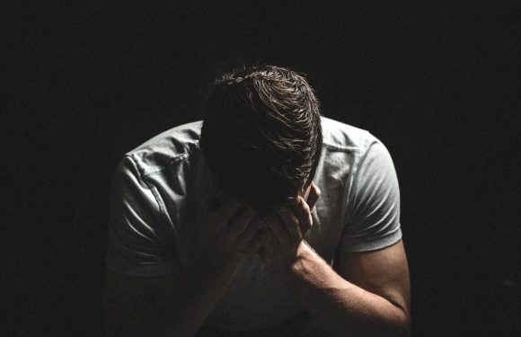 A man with short hair wearing a white shirt, hunched over with his hands covering his face, illuminated against a dark background, conveying distress or sorrow.