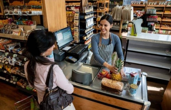 A smiling cashier with a pineapple and a loaf of bread at the checkout counter in a grocery store, interacting with a customer with a shopping bag.