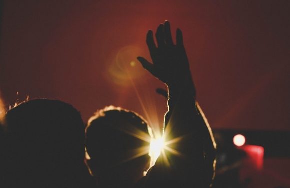 A person raising a hand with the sun shining behind them, creating a silhouette against a reddish sunset sky.