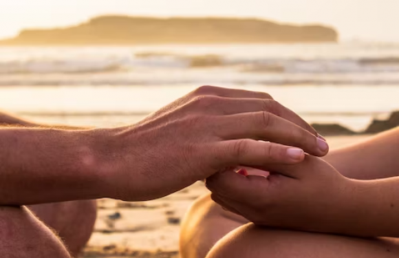 Hands gently holding each other on the beach during sunset with ocean waves and a distant landmass in the background.