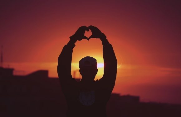 A person wearing a cap and a dark jacket is making a heart shape with their hands against a sunset background. The silhouette is dark with the bright sun behind.