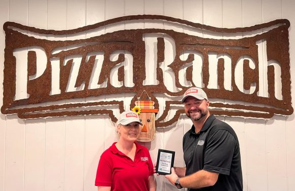 A man and woman wearing Pizza Ranch uniforms and hats stand in front of a large rustic Pizza Ranch sign. The man holds a framed award, and there is a decorative scarecrow face on the wall between them.
