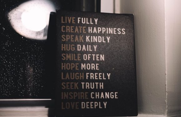 A black sign with motivational quotes in white and beige text, placed on a windowsill next to a window with raindrops on the glass.